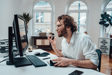 Portrait of a angry office worker with curly hairs he sits at table and raging on computer in...