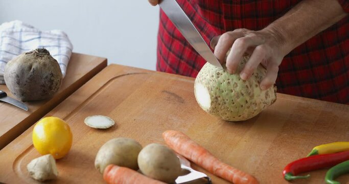 Vegetable soup preparing, cutting celery root into slices at cutting board using sharp knife, close up