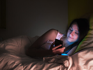 Young woman lying in bed checking social networks on her smartphone before going to sleep.