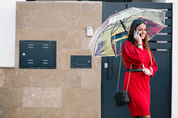 Portrait of a young blonde woman in a red dress talking on the mobile phone under an umbrella while...