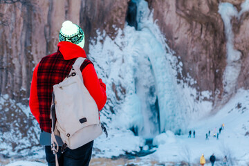 A tourist with a backpack on his back looks into the distance at the winter waterfall. Winter waterfall top view. 