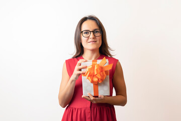 A young woman in a red dress and glasses holding a box with red ribbon, considering, twists a gift for the holiday. The girl looks at the new gift and smiles, becomes happy