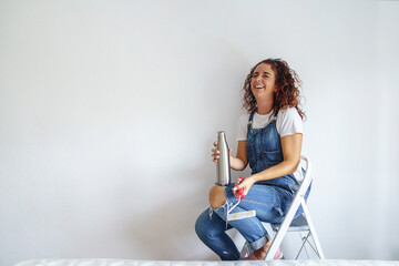 Horizontal view of caucasian woman resting sitting on a ladder after painting a wall at home. Young woman working with a paint roller drinking a refreshment after hard work in her new apartment.