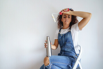 Horizontal view of caucasian woman exhausted resting on a ladder after painting a wall at home. Young woman working with a paint roller drinking a refreshment after hard work in her new apartment.