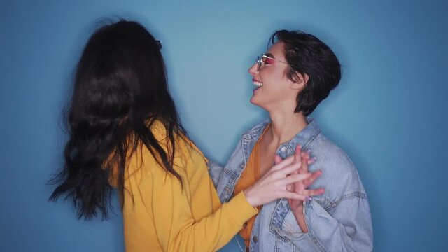 Two happy excited pretty stylish cool diverse gen z girls friends wearing glasses, trendy sunglasses and outfits having fun at hen night club party event, dancing together isolated on blue background.