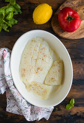 Raw halibut fish fillet, tomato, lemon, green basil and spices. White casserole on wooden rustic table, top view - 410474619