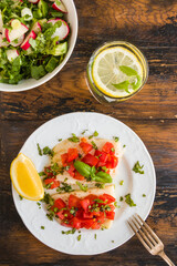 Halibut fish fillet baked with tomatoes on white plate. Salad with radish and cucumber in bowl. Wooden rustic table, top view - 410474672