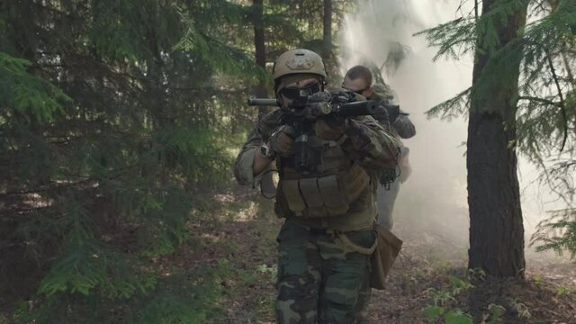 Tracking medium shot of soldiers in camouflage uniform armed with sniper rifles walking along forest through smoke bombs