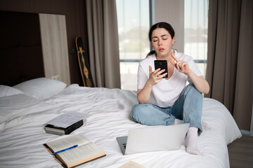 Young pretty woman is sitting on a bed and using a smartphone. Copy space. Books and laptop is nearby. The concept of online education and dependence on social networks