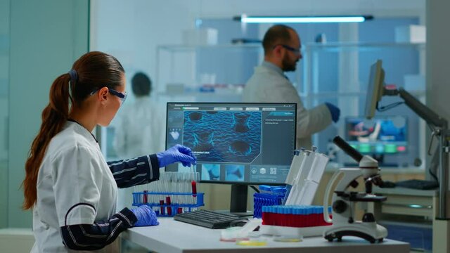 Chemistry doctor analysing blood sample and typing on computer while coworker opening door using digital computer with touch scan. Woman examining virus evolution using high tech and chemistry tools