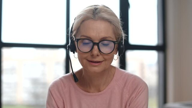 Helpful and friendly middle-aged woman wearing headset and stylish eyeglsses looks at the camera and smiles. Close-up headshot of mid-age female call-center employee, support officer on touch