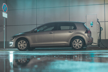 Side view of a modern electric car being charged on electric charging point or station. Night time neon style setting. Rain reflection on the ground.