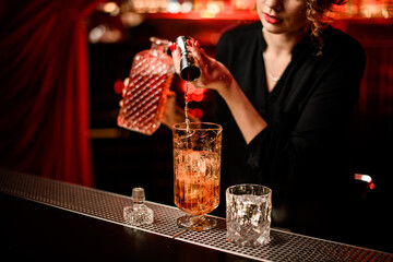 close-up of mixing cup with ice cubes into which woman bartender pours drink from jigger