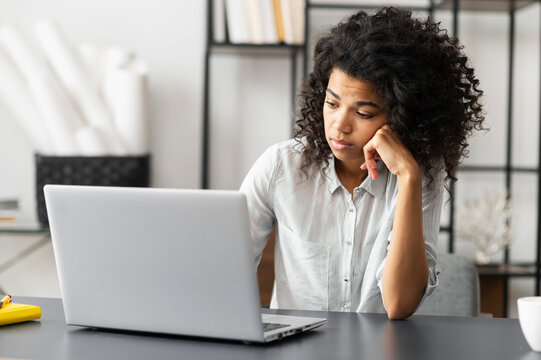 Young African American female trainee or an office worker with Afro hairstyle sitting at the desk, feeling bored and sleepy does not want to do homework or work on the project, unhappy with the task