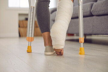 Close up view from behind the legs of a woman with a broken leg in plaster walking with crutches in the living room. Woman is undergoing rehabilitation at home. Concept of recovery after an accident.