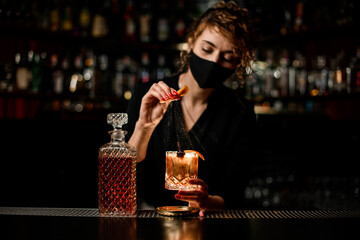 view on woman bartender in mask who holds glass of cocktail and sprinkles on it