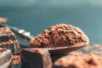 Brown cocoa powder in the spoon and chopped chocolate cubes on dark background with copy space