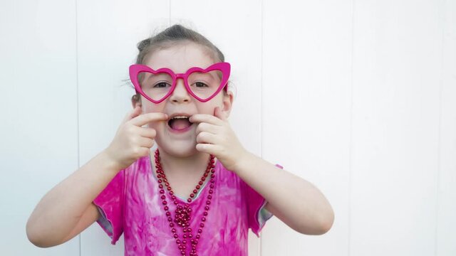 Positive dark hair and brown eyes girl in funny red heart glasses, beads and pink dress making funny silly faces at the camera. Celebrating Valentines day. White wall background