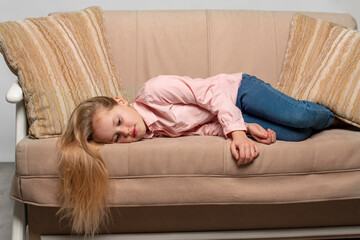 A little blonde girl with long hair wearing pink shirt and jeans  lies on the couch and is sad.