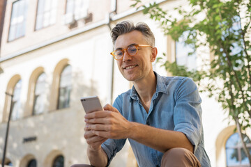 Portrait of happy freelancer receive payment sitting in park. Handsome mature businessman holding smartphone, communication, reading news