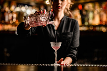 Close-up view of beautiful female bartender pouring cocktail from mixing cup into crystal glass