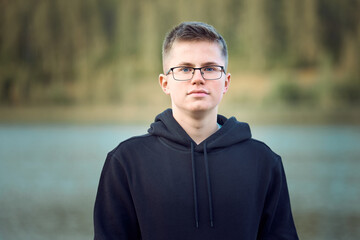 Portrait of serious teenager boy with glasses in black hoodie on the background of lake and forest in blur.