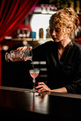 view of beautiful female bartender pouring cocktail from mixing cup into crystal glass