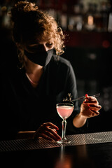 woman bartender in mask carefully decorates glass of pink cocktail with flower sprig