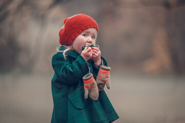 Funny little girl in red hat and greet coat is eating a candy in autumn park. Image with selective focus, toning and noise effect