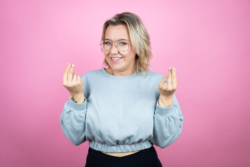 Young caucasian woman wearing sweatshirt over pink background doing money gesture with hands, asking for salary payment, millionaire business