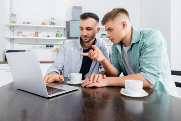 homosexual man pointing with finger and looking at laptop near husband and cups on table