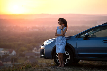 Happy young woman driver in blue dress enjoying warm summer evening standing beside her car. Travelling and vacation concept.