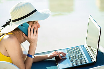 Young woman on beach chair at swimming pool working on computer laptop and talking on sell phone in...