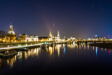 Fototapeta na wymiar Night cityscape view of historic buildings with reflections in Elbe river in the center of Dresden (Germany) and starry dark sky with Neowise comet with light tail.