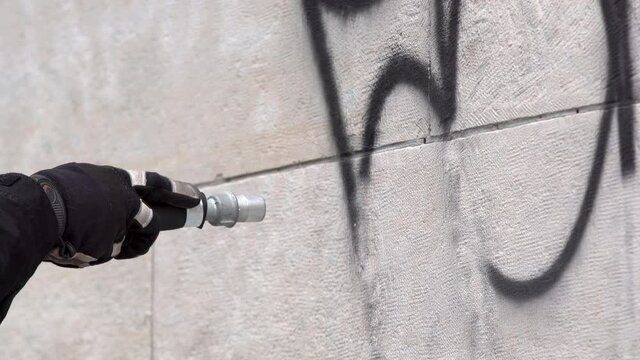 Worker cleans graffiti from a public building wall using pressure sand blaster.