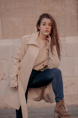 Beautiful blonde girl in a brown jacket with a blue trousers while her hands are touching into her face on a background of architecture.