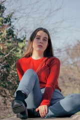 Obraz premium Close up picture of young thoughtful woman in red jersey and black high heels looking in front at the camera in a winter sunny day while sitting on a bench in side of a park.