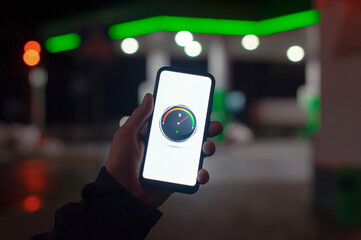 A man holds a smartphone with a digital fuel meter on the screen against the background of a night...