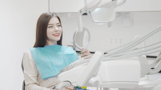 Cheerful young woman smiling to the camera sitting in dental chair. Attractive female patient looking in the mirror after teeth care treatment at dental clinic