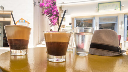 two ice coffees and two glasses of water and a hat in focus on a table with a blurred background of sunny white houses on Kythira, Greece