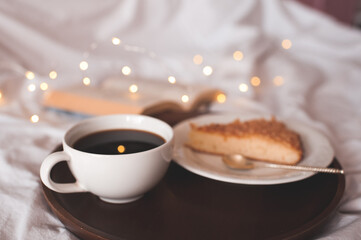 Cup of fresh coffee with tasty pie on white plate with open book in bed over glosing lights close up. Good morning. Breakfast time.