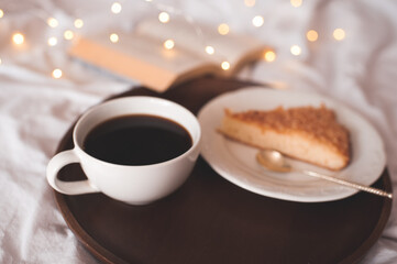 Cup of fresh coffee with tasty pie on white plate with open book in bed over glosing lights close up. Good morning. Breakfast time.