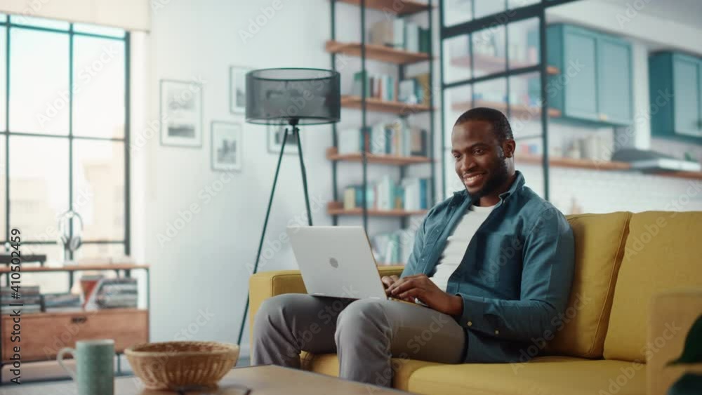 Poster handsome black african american man working on laptop computer while sitting on a sofa in cozy livin