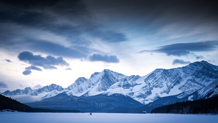 Obraz na płótnie Canvas Distant fishermen stand in the middle of the frozen Kananaskis Lake surrounded by the Canadian Rocky Mountains in Alberta Canada.