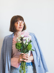 Young woman wearing blue jacket and pink shirt holding beautiful pastel peony bouquet on light gray background