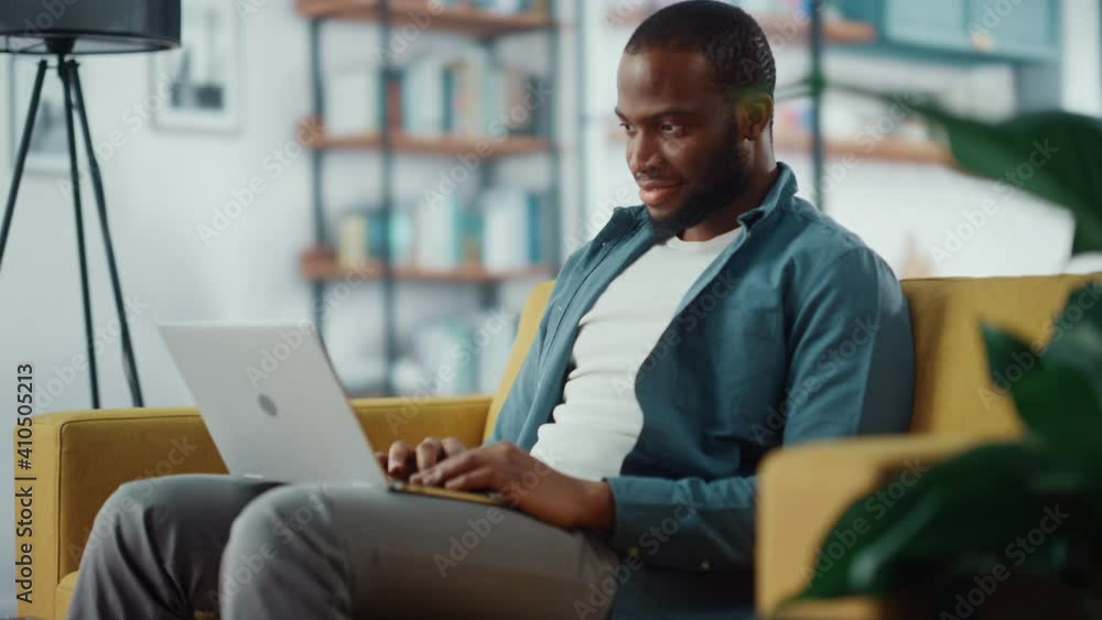 Poster handsome black african american man working on laptop computer while sitting on a sofa in cozy livin