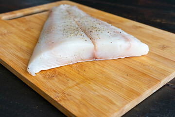 Raw Halibut Fillet on a Bamboo Cutting Board: Uncooked white fish fillet seasoned with salt and...