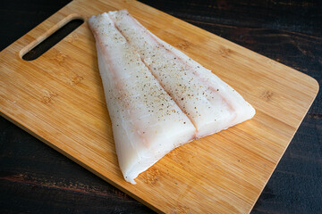 Raw Halibut Fillet on a Bamboo Cutting Board: Uncooked white fish fillet seasoned with salt and...