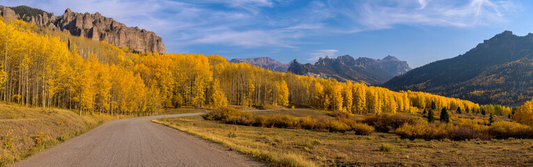 Autumn Golden Valley - A panoramic Autumn evening view of Owl Creek Pass Road winding through a dense golden aspen grove surrounded by the rugged mountain ridges, near Ridgway, Colorado, USA.