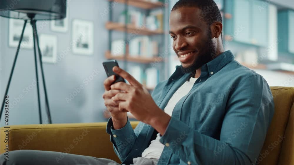 Poster handsome black african american man using smartphone while sitting on a sofa in cozy living room. fr